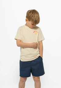 Beige bomull t-shirt med texten "Time Flies" och ett snäcka-motiv på bröstet, tillsammans med marinblå shorts, stående pojke med händerna i fickorna.