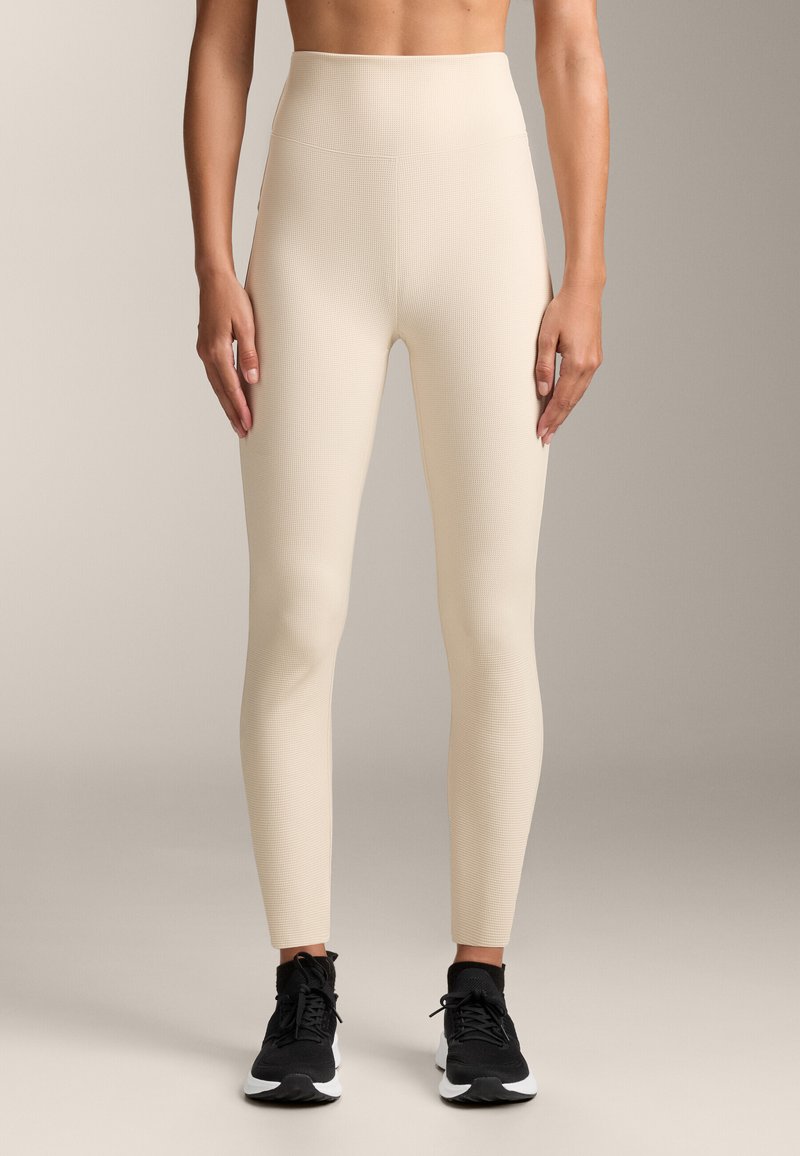 Visoko pasne bež leggins z teksturiranim vzorcem, oblikovano prileganje in gladko tkanino. V kombinaciji s črnimi športnimi čevlji.