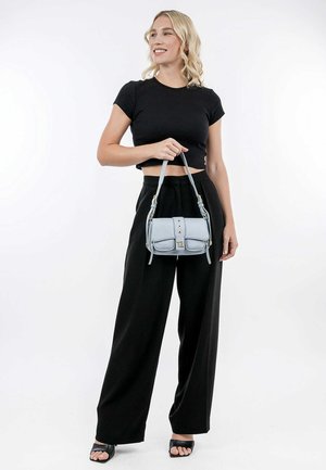 Light blue structured handbag with dual front pockets, silver hardware, and a top handle, held by a person in black cropped top and wide pants.