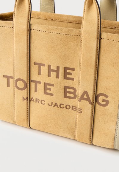 Τσάντα tote από ταμπά σουέντ με χοντρές λαβές και την επιγραφή "THE TOTE BAG MARC JACOBS" σε μεγάλα καφέ γράμματα στο μπροστινό μέρος.