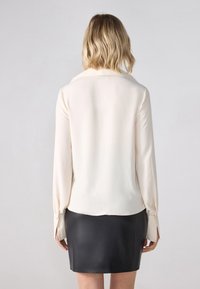 Kremfarget bluse med lange ermer og knapper på mansjett, kombinert med et svart skinn miniskjørt. Glatt tekstur og skreddersydd design.