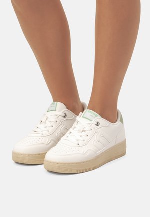 Zapatillas blancas de cuero con puntera perforada, acento verde en el talón y suela de goma beige. Los cordones y ojales son estándar.