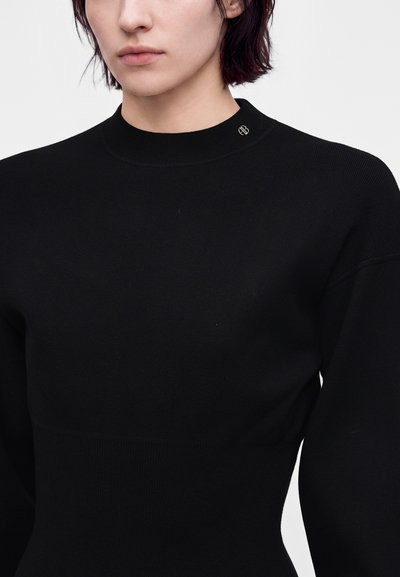 Femme aux cheveux courts et foncés portant un haut noir près du corps à manches longues avec un petit logo circulaire près de la clavicule sur un fond uni.