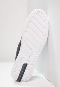 Baskets noires avec une semelle en caoutchouc blanche présentant des rainures texturées. Le côté affiche un logo et une bande accentuée bleu marine.