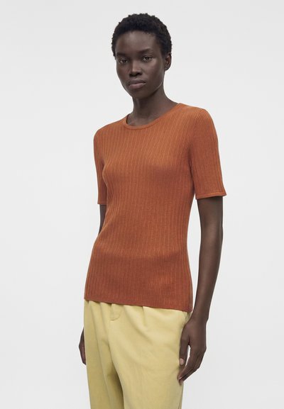 Γυναίκα με κοντά σγουρά μαλλιά που φορά εφαρμοστό ριμπ μπλουζάκι με κοντά μανίκια σε σκουριά απόχρωση και χαλαρά κίτρινα παντελόνια πάνω σε απλό φόντο.