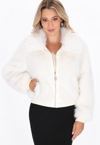 Frau in einer weißen, flauschigen Zip-Jacke mit großem Kragen, goldenen Creolen und einem schwarzen Rock, die mit einer Hand in der Tasche posiert.