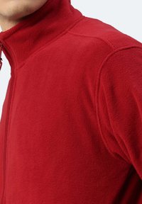 Giacca in fleece rossa con colletto alto, dotata di zip a tutta lunghezza e cuciture visibili. Tessuto morbido e colore solido, senza motivi o dettagli.