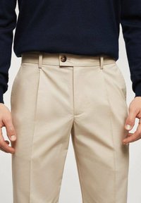 Pantaloni beige in tessuto liscio, con vestibilità affusolata, due pieghe frontali e chiusura con bottone marrone in vita.