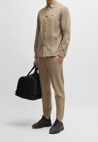 Veste légère beige avec des boutons noirs et un logo, assortie à un pantalon tapered et des baskets noires, tenant un sac noir.
