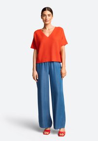 Rode T-shirt met korte mouwen en V-hals, gecombineerd met blauwe wijde broeken en rode haksandalen. De stof lijkt zacht en heeft een ontspannen pasvorm.