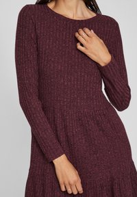 Femme portant une robe maroon en tricot côtelé à manches longues et décolleté rond, montrant des mains avec de fines bagues en argent aux doigts.