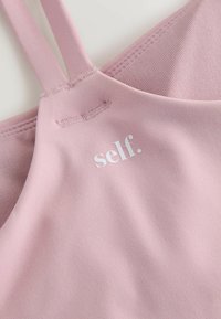 Hele roosa spordirinnahoidja reguleeritavate rihmadega, valmistatud tekstuurse kangaga, millel on valge trükitud logo "self." kaeluse juures.