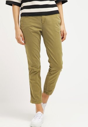 Vrouw die een korte olijfgroene broek, witte sneakers en een zwart-wit gestreepte top draagt, staand tegen een effen witte achtergrond.