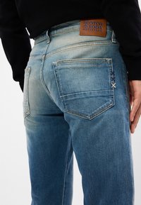 Απαλές μπλε τζιν παντελόνες διαθέτουν ξεθωριασμένο σχέδιο, πέντε τσέπες και μια καφέ δερμάτινη ετικέτα με "Scotch & Soda Amsterdam" στη μέση.