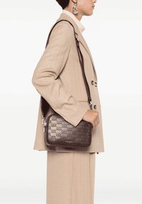 Borsa a tracolla in pelle marrone con texture e logo MK ripetuto, forma rettangolare, tracolla regolabile, abbinata a un cappotto beige su misura.