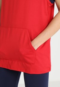 Person med en rød ærmeløs top med en frontlomme, hånden stukket indeni, parret med mørkeblå bukser.