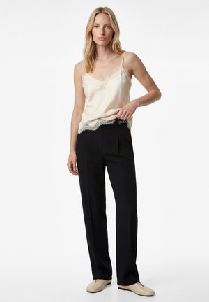 Donna in piedi che indossa un camicione di raso color crema con bordi in pizzo, pantaloni neri a vita alta con dettaglio cintura e scarpe beige slip-on su sfondo bianco.