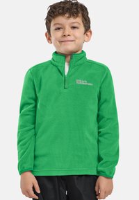 Ragazzo con capelli ricci corti che indossa un pullover in pile verde brillante con mezzo zip e pantaloni neri, in piedi su uno sfondo uniforme.