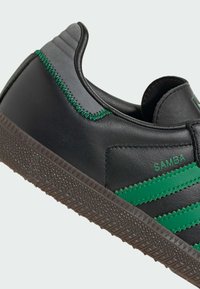 Black leather sneaker with green accents, featuring three stripes, textured brown rubber sole, and contrasting stitching along the heel.