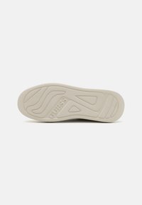 Hellbeige Sneaker-Sohle mit strukturiertem Oberflächen- und Wellenmuster. Auf der Seite befindet sich der geprägte Markenname "GUESS".