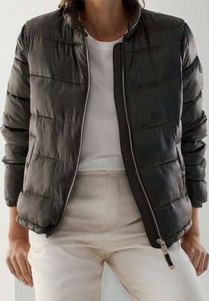Personne portant une veste matelassée noire ouverte sur une chemise blanche et un pantalon beige clair, debout les mains le long du corps.