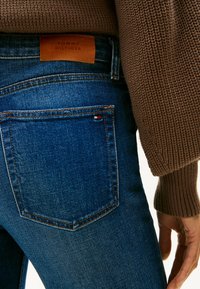 Jeans en denim bleu foncé avec un pull texturé marron. Présente une étiquette de marque en cuir et un petit accent tricolore sur la poche arrière.