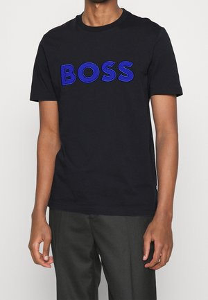 T-shirt noir à manches courtes avec le texte "BOSS" en bleu majuscules porté par une personne portant un pantalon gris foncé, debout devant un fond clair uni.
