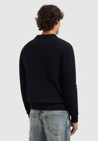 Zwarte gebreide trui met een gestructureerde afwerking, voorzien van een kraag en ribgebreide manchetten, gecombineerd met lichtblauwe denim jeans.