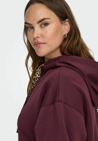 Burgundijas krāsas džemperis ar leopardu raksta akcentiem uz kapuces, ar mīkstu tekstūru un brīvu piegriezumu, redzams no sānu skatu.