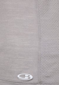 Top atletico grigio chiaro con texture liscia e tessuto a fantasia su un lato; include un logo bianco nell'angolo inferiore.