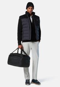 Schwarze gesteppte Weste über einem schwarzen Strickpullover, kombiniert mit hellen Hosen und schwarzen Turnschuhen. Eine schwarze Reisetasche mit geprägt-Logo und Griffen.