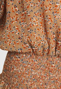 Blusa floreale con uno sfondo marrone e motivi arancioni e blu, con arricciatura elastica in vita e tessuto testurizzato.