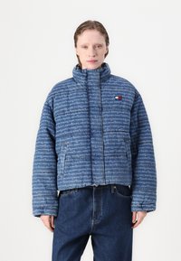 Blauwe pufferjack met een hoge kraag, voorzien van een volledig logo-patroon en voorzakken, gecombineerd met losse blauwe jeans.