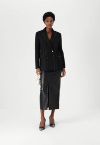 Blazer nero doppiopetto con cuciture bianche, abbinato a una gonna a matita in pelle nera con spacco frontale e tacchi neri.