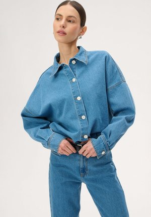 Kobieta poprawiająca czarny pasek, ubrana w za dużą jasnoniebieską kurtkę dżinsową i pasujące dżinsy, z włosami związanymi do tyłu i minimalnym makijażem.