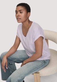 Licht lavendelkleurig V-hals T-shirt met opgerolde korte mouwen, gecombineerd met lichtblauwe flared jeans, zittend op een gebogen crèmekleurige stoel.