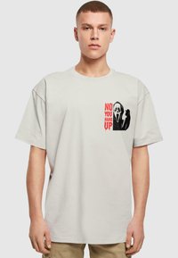 Lys grå kortærmet t-shirt med et sort og rødt grafisk print af et spøgelsesansigt og teksten "NEJ, DU LÆG PÅ" på brystet.