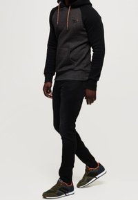 Felpa con cappuccio nera e grigia con coulisse, abbinata a pantaloni neri slim-fit e sneaker olive con accenti arancioni.