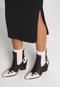 Svarta läderankelstövlar med vita blommönster, spetsiga tår och en kraftig klack. Har ribbade paneler och en kontrasterande grön flik.