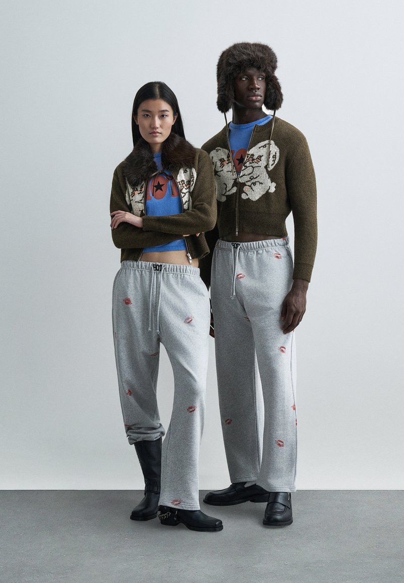 Δύο μοντέλα φορούν συντονισμένα ρούχα. Το ένα φοράει καφέ κεντημένο πουλόβερ με ψεύτικη γούνα και μπλε μπλουζάκι με γραφικό σχέδιο· το άλλο φοράει γκρι παντελόνι φόρμας με αποτυπωμένα χείλη και μπότες.