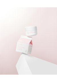 Weißes Gefäß mit "White in Whipping Cream" in rosa Schrift, zusammen mit einem rosa-weißen Karton, auf einer weißen geometrischen Oberfläche. Minimalistisches Design.