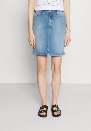 Donna che indossa una gonna di jeans blu a metà coscia, una maglia bianca infilata dentro e sandali neri con due grandi fibbie, in piedi contro uno sfondo neutro.