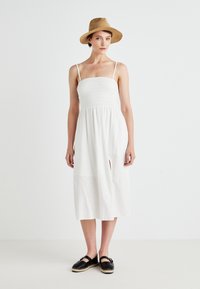Vestido midi de algodón blanco con corsé con fruncido, tirantes finos y abertura lateral. Combinado con zapatos estilo espadrille negros y un sombrero de paja.
