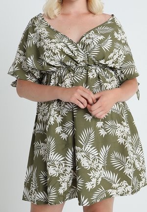 Kvinde iført grøn off-shoulder kjole med hvidt tropisk blad mønster, hænder samlet ved taljen, lyst baggrund.