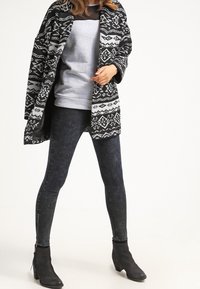 Zwart-wit patroon oversized jas over een grijze sweater, gecombineerd met donkergewassen skinny jeans en zwarte enkellaarsjes.