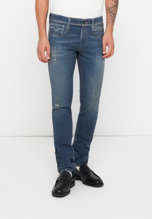 Mężczyzna ubrany w obcisłe niebieskie dżinsy z delikatnym przetarciem i czarne skórzane loafersy, stojący na tle jednolitego białego tła.