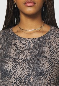 Elegant top s hadem hadovitého vzoru v černé a béžové barvě, s kulatým výstřihem, doplněný o vrstvené náhrdelníky ve zlatém tónu a kruhové náušnice.