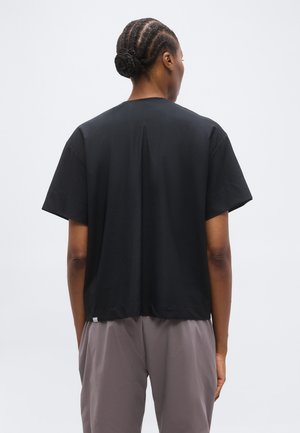 Person with braided hair in a black short-sleeve shirt and loose gray pants, standing facing away against a plain light background.