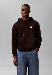 Muškarac odjeven u crnu Calvin Klein hoodicu s prednjim džepom i plave traperice, stoji s jednom rukom u džepu na sivoj pozadini.