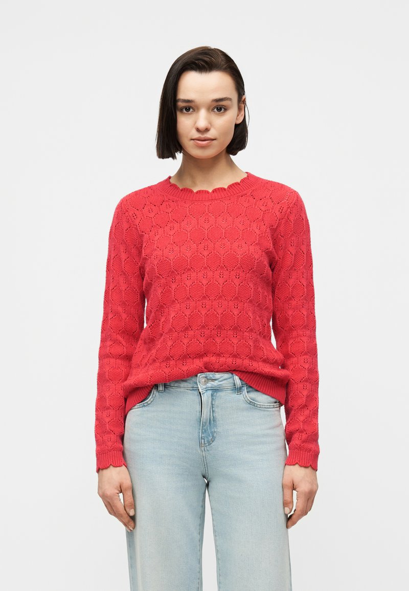 Jeune femme aux cheveux longs et lisses et foncés, portant un pull à manches longues rouge vif à motifs et un jean bleu clair, debout devant un fond uni.